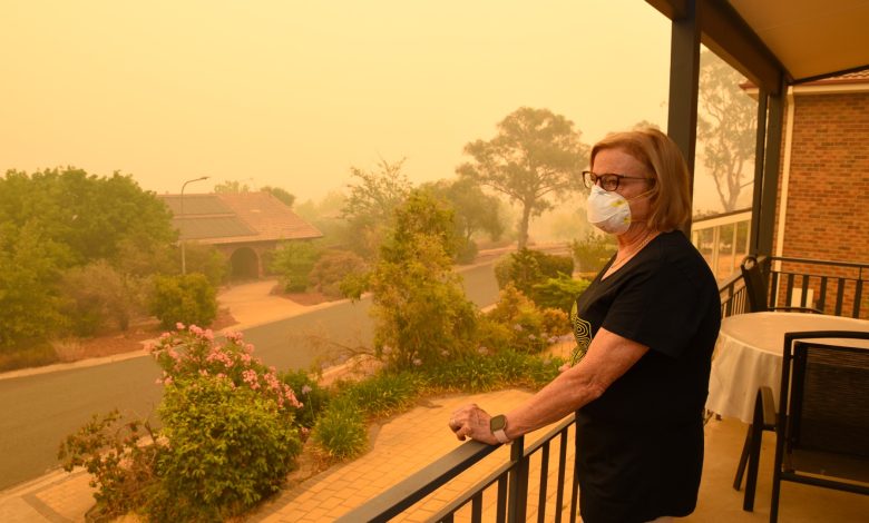 Seorang wanita dalam gambar berdiri di balkonnya memandang ke kejauhan. Langit diwarnai oranye/kuning dan dia mengenakan masker pernapasan.