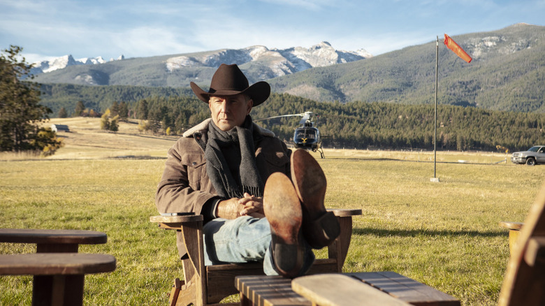 Kevin Costner sebagai John Dutton duduk dengan kakinya bersilang dan helikopter di latar belakang di Yellowstone