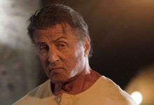 Rambo (Sylvester Stallone) terlihat di sebelah kanan dengan ekspresi yang serius dan marah, mengenakan kemeja kancing krem, saat cahaya kabur bersinar di latar belakang di Rambo: darah terakhir