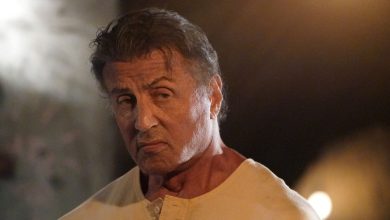 Rambo (Sylvester Stallone) terlihat di sebelah kanan dengan ekspresi yang serius dan marah, mengenakan kemeja kancing krem, saat cahaya kabur bersinar di latar belakang di Rambo: darah terakhir