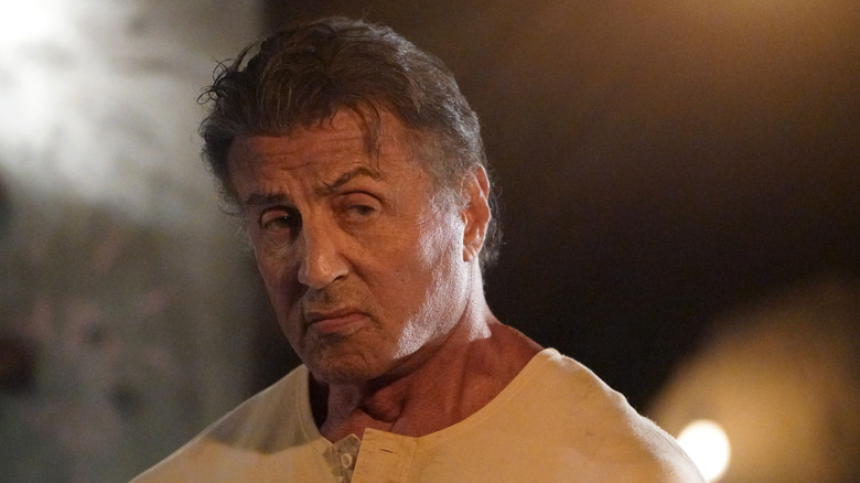 Rambo (Sylvester Stallone) terlihat di sebelah kanan dengan ekspresi yang serius dan marah, mengenakan kemeja kancing krem, saat cahaya kabur bersinar di latar belakang di Rambo: darah terakhir