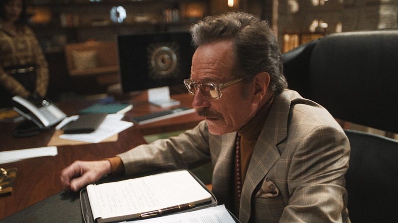 Bryan Cranston sebagai Griffin Mill duduk di mejanya di studio