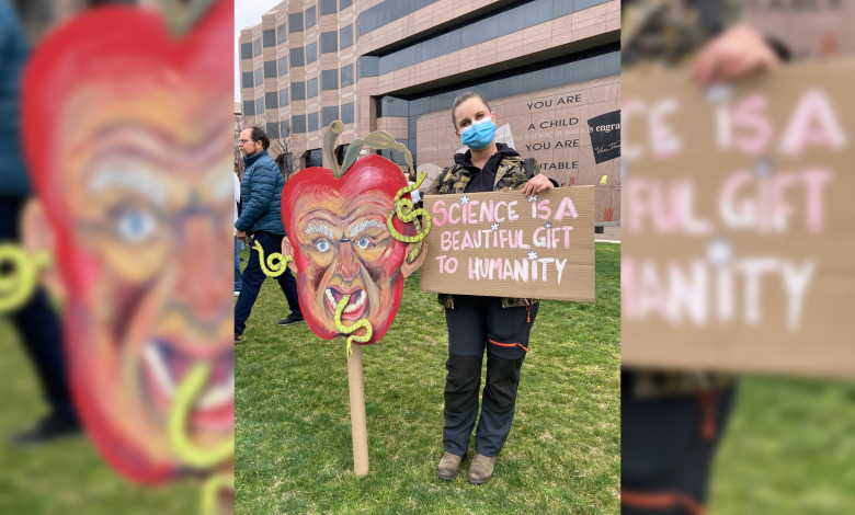 Tembakan poster dan pemrotes di Stand Up For Science Rally di Raleigh; Seorang wanita berdiri dengan dua poster, salah satunya adalah kepala RFK Jr yang sangat besar sebagai apel busuk dengan cacing