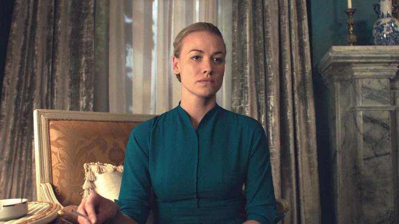 Serena (Yvonne Strahovski) dengan ekspresi khidmat dan fokus, mengenakan gaun teal konservatif untuk istri dalam kisah pelayan