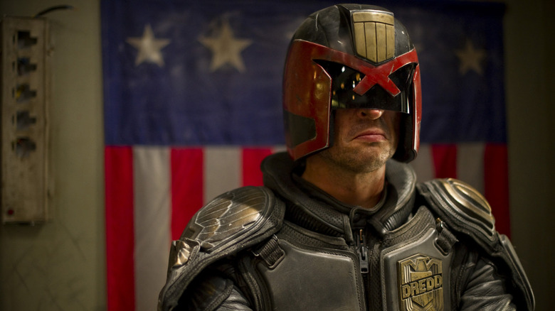 Karl Urban sebagai Hakim Dredd berdiri di depan bendera di Dredd