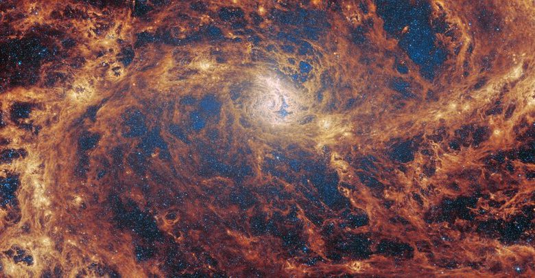 Pandangan close-up dari galaksi spiral yang dilarang. Dua lengan spiral mencapai horizontal jauh dari inti di tengah, menggabungkan ke jaringan gas dan debu yang luas yang mengisi gambar. Bahan ini bersinar oranye paling terang di sepanjang jalan lengan, dan merah lebih gelap di seluruh galaksi. Melalui banyak celah di debu, bintang -bintang kecil yang tak terhitung dapat dilihat, paling padat di sekitar inti