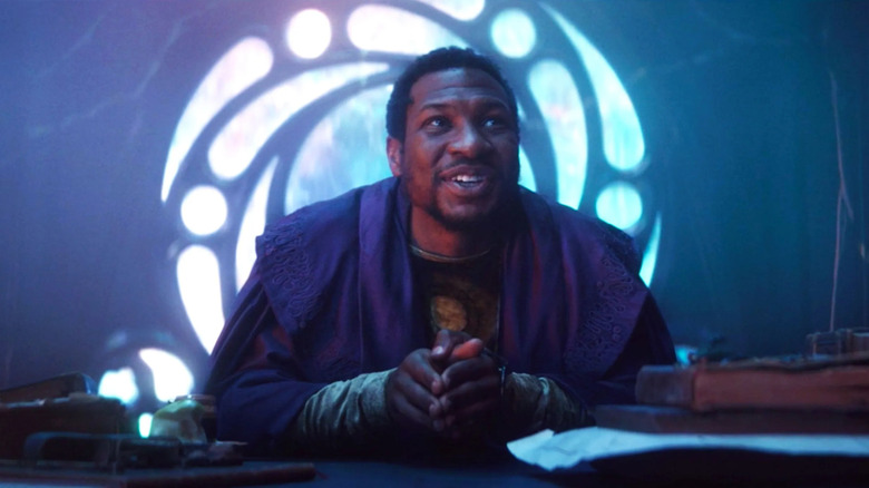 Jonathan Majors 'Dia tetap, mengenakan jubah ungu, bersandar ke depan dan tersenyum pada seseorang di luar layar di benteng pada akhir waktu di Loki musim 1
