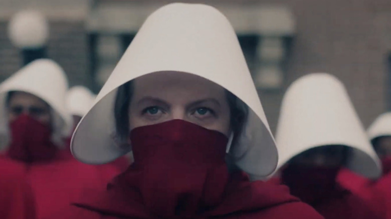 Juni (Elisabeth Moss) memakai jubah pelayan merah, kap putih, dan wajah merah yang menutupi ditarik ke hidungnya, berdiri di antara pelayan tangan lainnya dan menatap ke depan dengan tatapan yang kuat dalam kisah pelayan handmaid itu