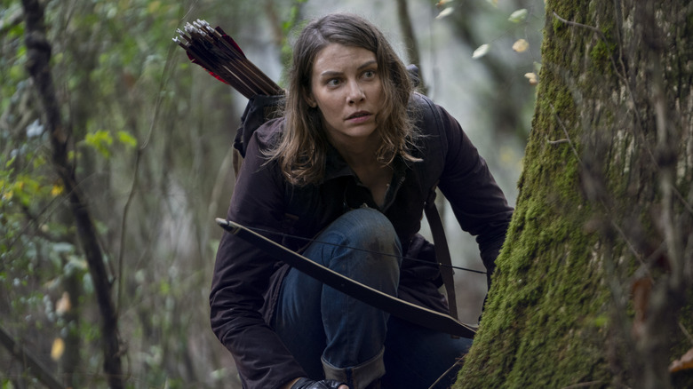 Lauren Cohan sebagai Maggie Greene berlutut di tepi pohon dengan busur dan panahnya di jalan mati