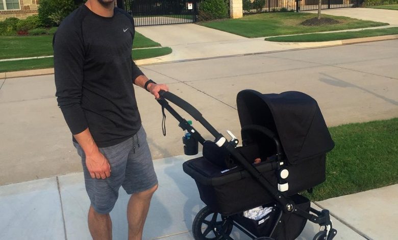 Justin Tucker mengenakan topi baseball hitam, kemeja hitam, dan celana pendek abu -abu, tersenyum sambil menggendong kereta dorong bayi