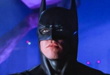 Val Kilmer sebagai Batman dengan kostum menantikan dengan latar belakang biru di belakangnya
