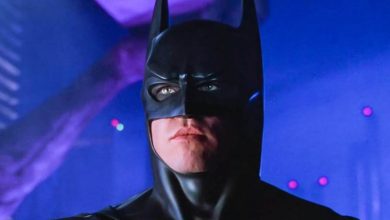 Val Kilmer sebagai Batman dengan kostum menantikan dengan latar belakang biru di belakangnya