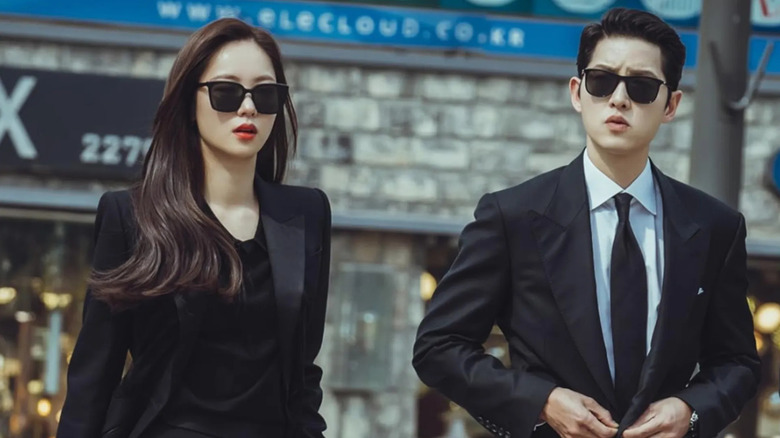 Hong Cha-Young (Jeon Yeo-Been) berjalan di samping Vincenzo Cassano (Song Joong-ki), dengan pakaian gelap dan kacamata hitam, di Vincenzo